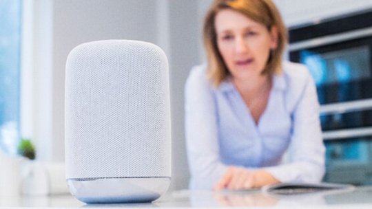 Une enceinte intelligente moderne est placée bien en vue sur un comptoir de cuisine blanc. À l'arrière-plan, une personne se penche en avant, suggérant une interaction ou une observation de l'appareil.