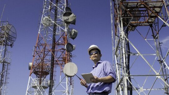 Une personne portant un casque de sécurité et tenant une tablette se tient parmi plusieurs grandes tours de télécommunications. Le cadre est en extérieur sous un ciel bleu clair, avec diverses antennes et paraboles visibles sur les tours.