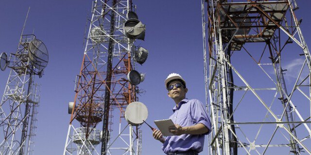 Une personne portant un casque de chantier et tenant une tablette se tient parmi plusieurs grandes tours de télécommunications. Le cadre est en plein air sous un ciel bleu clair, avec diverses antennes et paraboles visibles sur les tours.