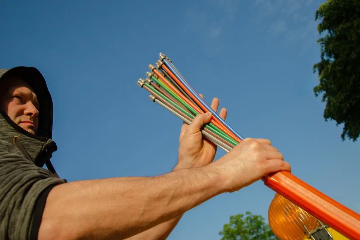 A person is holding a bundle of colorful metal rods, possibly tent poles or stakes, against a clear blue sky.