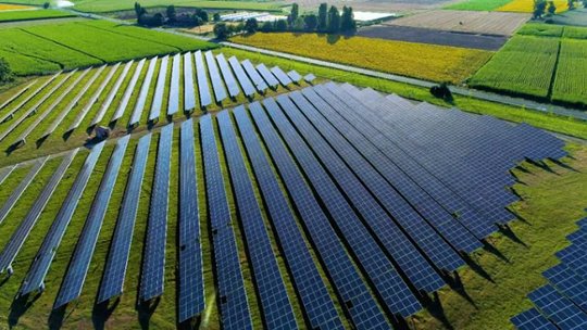 A wide aerial view shows an extensive solar panel installation set in a rural area surrounded by green fields. The solar panels are arranged in neat, parallel rows, capturing sunlight for renewable energy generation. The landscape is lush and vibrant, with clear skies and distant trees visible.