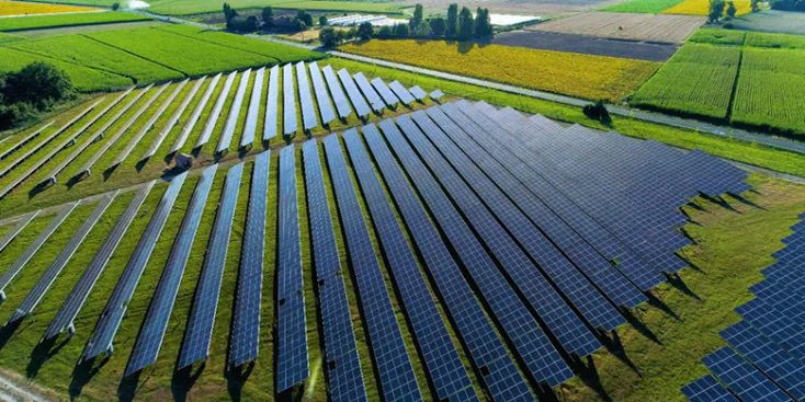 A wide aerial view shows an extensive solar panel installation set in a rural area surrounded by green fields. The solar panels are arranged in neat, parallel rows, capturing sunlight for renewable energy generation. The landscape is lush and vibrant, with clear skies and distant trees visible.