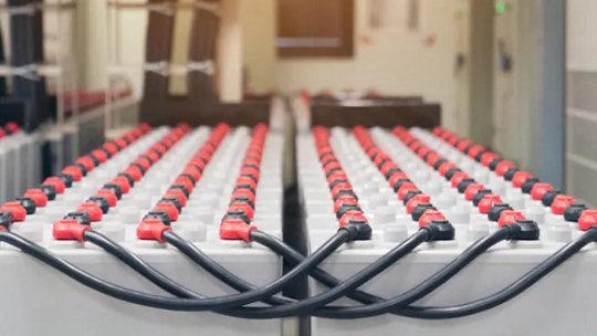 Multiple rows of large industrial batteries connected by thick black cables. The batteries are arranged in a clean, well-lit facility. Red and black connectors are visible on each battery.