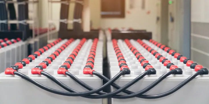 Multiple rows of large industrial batteries connected by thick black cables. The batteries are arranged in a clean, well-lit facility. Red and black connectors are visible on each battery.
