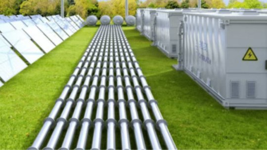 A solar energy installation featuring rows of solar panels on the left and multiple metallic pipes or power cables running through the center. On the right, there are large industrial containers with a visible high voltage warning symbol. The setting is outdoors on a grassy field with trees in the background, under daylight conditions.
