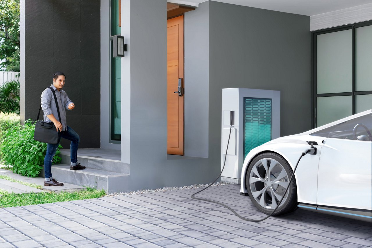 A man carrying a black bag approaches the entrance of a modern house. In the driveway, a white electric vehicle is plugged into a charging station.