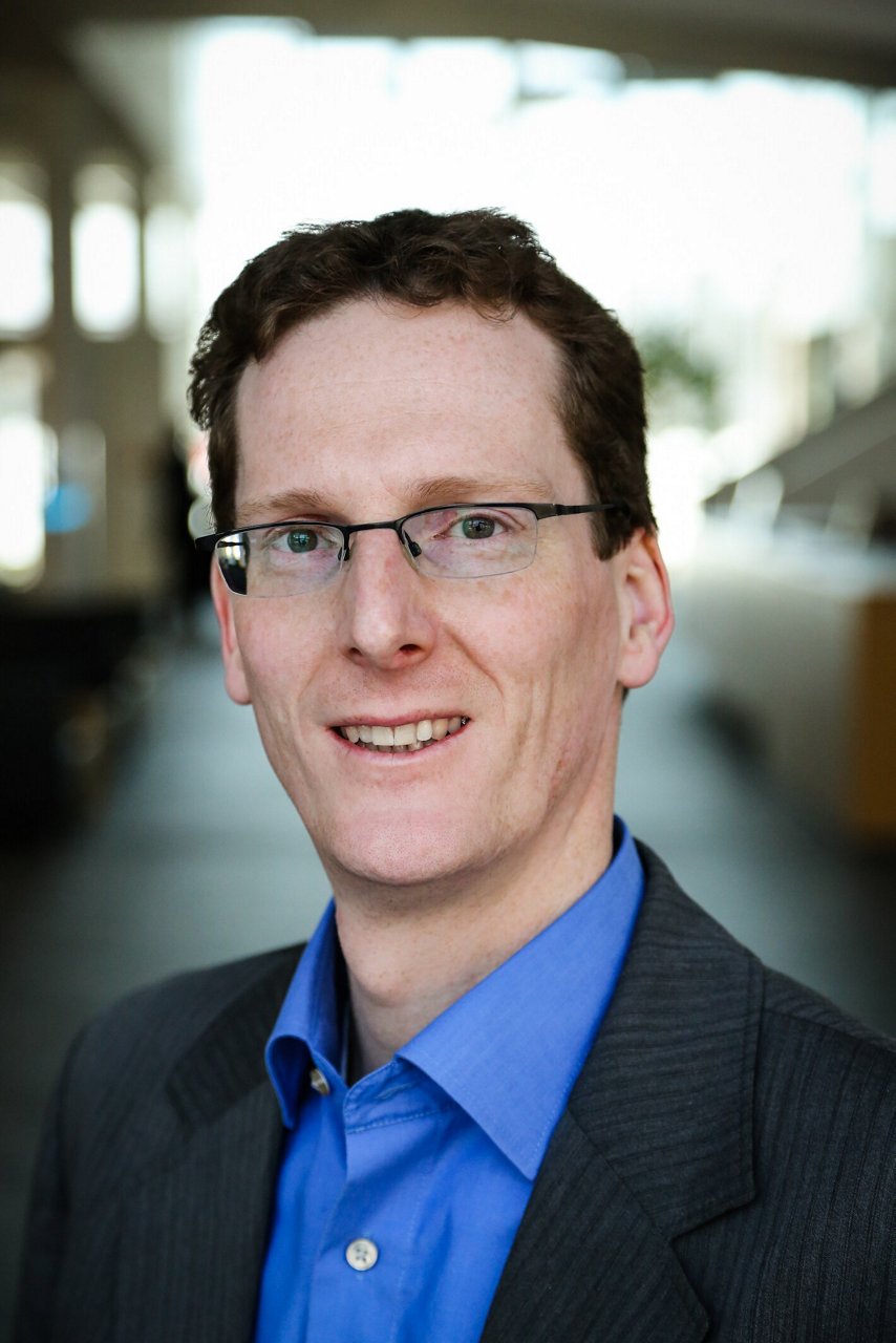 A man dressed in business attire stands in a contemporary indoor setting. The subject is wearing a blue collared shirt and a dark pinstripe suit jacket.