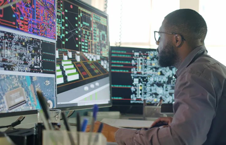 A person is seated at a desk, working on multiple large monitors displaying complex circuit board and electronic schematic diagrams. The workspace is filled with technical visuals, including detailed layouts and wiring patterns.