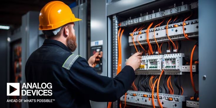 Un ingénieur masculin portant un casque de protection jaune examine un panneau de commande électrique ouvert rempli de câbles orange et de disjoncteurs. La scène est bien éclairée, mettant en évidence la complexité du câblage et de l'équipement. Le logo Analog Devices et la phrase 'AHEAD OF WHAT'S POSSIBLE™' sont clairement visibles dans le coin inférieur gauche.