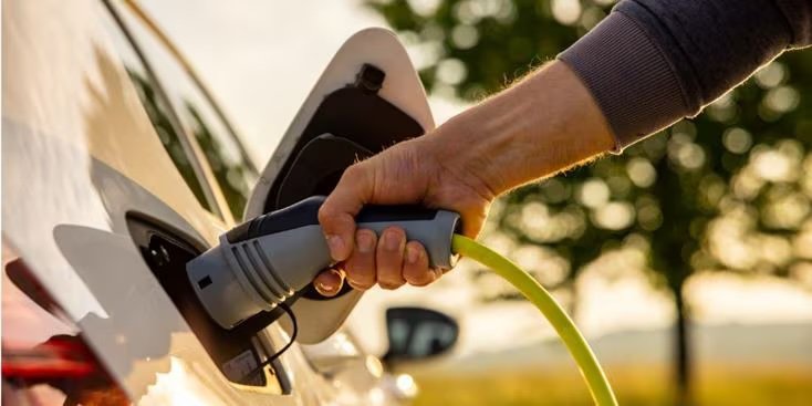 A man inserts a power cord into an electric car for charging in nature