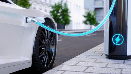 A white electric vehicle is plugged into a modern charging station on a city sidewalk. The image highlights the glowing charging cable and the station's illuminated power symbol.