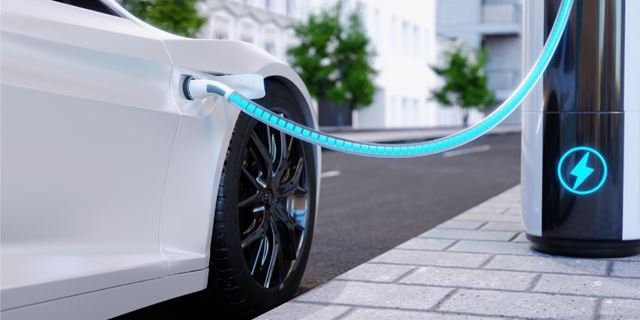 A white electric vehicle is plugged into a modern charging station on a city sidewalk. The image highlights the glowing charging cable and the station's illuminated power symbol.