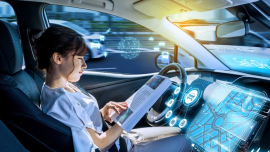 A woman sits in the driver's seat of a modern car, reading a magazine while the vehicle operates autonomously. 