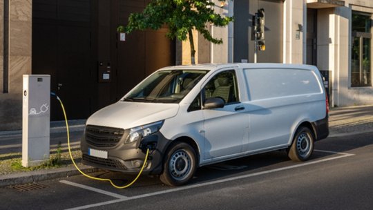 A white electric delivery van is parked on a city street, connected to a public charging station. 