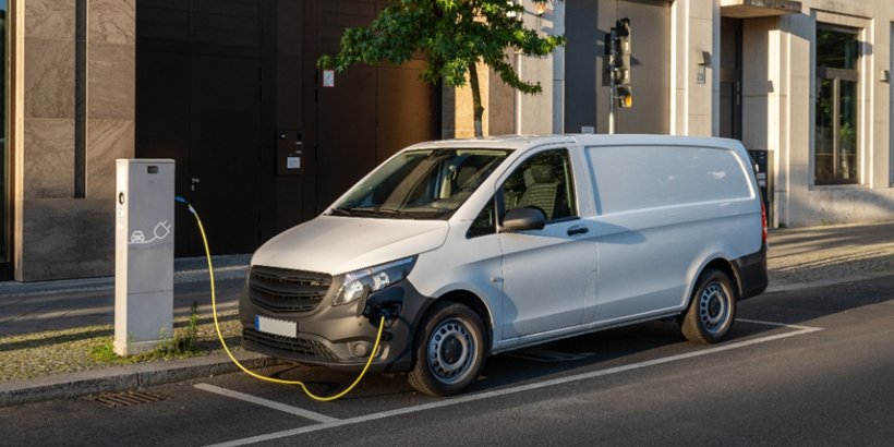 A white electric delivery van is parked on a city street, connected to a public charging station. 