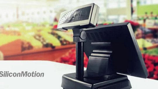 Un terminal point de vente de marque SiliconMotion est affiché en premier plan. L'appareil dispose d'un écran orienté vers le client et est situé dans un environnement de supermarché ou épicerie bien éclairé. L'arrière-plan est flou, mettant en valeur des produits frais et des étagères de magasin. Le logo et le nom SiliconMotion sont clairement visibles dans le coin inférieur gauche.