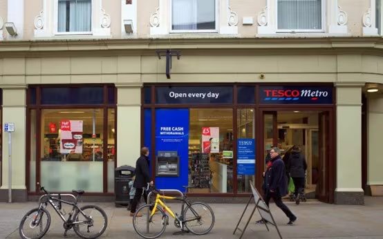 The image shows the exterior of a Tesco Metro store with a clearly visible ATM labeled 'Free Cash Withdrawals.' Two bicycles are parked in front, and two adults are walking past the entrance. The store signage and window displays are prominent, and the setting appears to be an urban street during daytime.