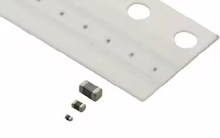Three small surface mount electronic components placed next to a white ruler or measurement guide. The ruler features two large circular holes and several smaller markings for scale reference. 