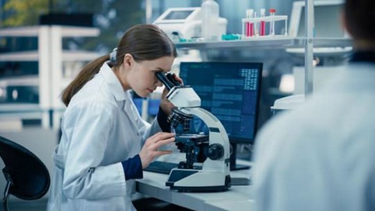 Uno scienziato in camice bianco osserva attentamente un campione attraverso un microscopio in un ambiente di laboratorio moderno. Lo spazio di lavoro presenta un monitor del computer che visualizza dati, vetreria da laboratorio e attrezzature scientifiche.
