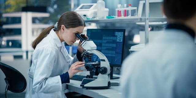 Un scientifique, vêtu d'une blouse blanche de laboratoire, observe attentivement un échantillon au microscope dans un laboratoire moderne. L'espace de travail comprend un écran d'ordinateur affichant des données, de la verrerie de laboratoire et des équipements scientifiques.