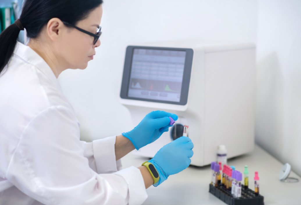 A laboratory technician wearing blue gloves handles a blood sample vial in a clinical setting. The technician is seated at a workstation with a modern hematology analyzer displaying data on its screen.