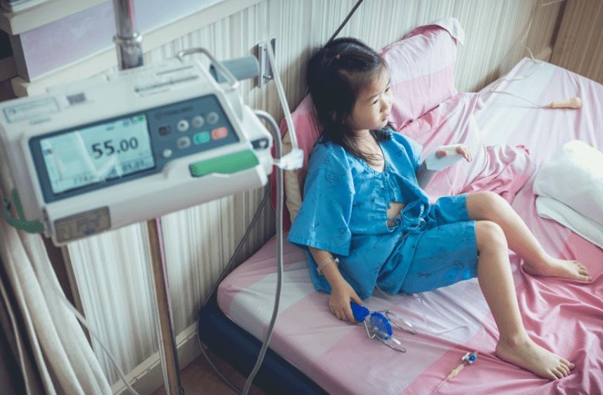 A young child is seated on a hospital bed, wearing a blue patient gown. An infusion pump displaying '55.00' is visible in the foreground.