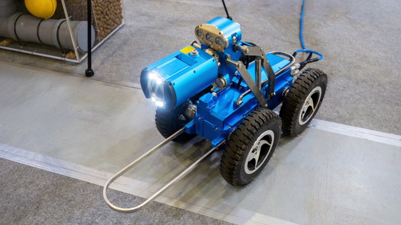 A modern agricultural robot navigates between rows of lush green plants inside a greenhouse. The robot is equipped with wheels and visible wiring, designed for automated crop management. The scene is brightly lit with natural sunlight, highlighting the advanced technology and healthy vegetation. No visible text or numeric values are present on the robot or in the environment.