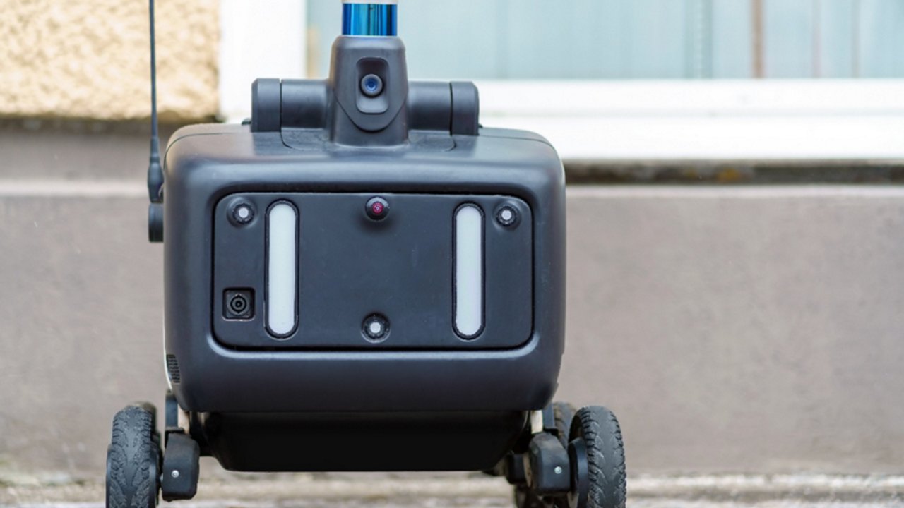 A modern agricultural robot navigates between rows of lush green plants inside a greenhouse. The robot is equipped with wheels and visible wiring, designed for automated crop management. The scene is brightly lit with natural sunlight, highlighting the advanced technology and healthy vegetation. No visible text or numeric values are present on the robot or in the environment.