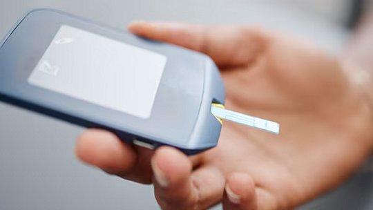 특히 대형 이미지에서, 한 사람이 테스트 스트립이 삽입된 혈당 측정기를 들고 있는 모습을 나타냅니다. 기기는 파란색이며 흰색 라벨 영역이 비어 있고, 배경에서 사람의 손이 보입니다.