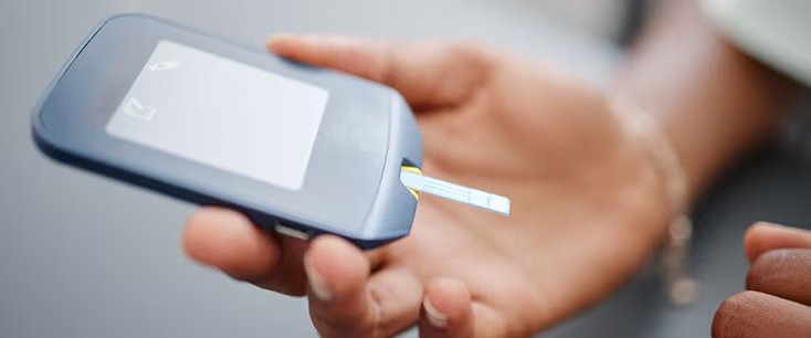 근접 촬영 이미지에는 테스트 스트립이 삽입된 혈당 측정기를 들고 있는 사람이 보입니다. 장치는 파란색이며, 흰색 라벨 공백 영역이 있습니다. 배경에는 그 사람의 손이 보입니다.