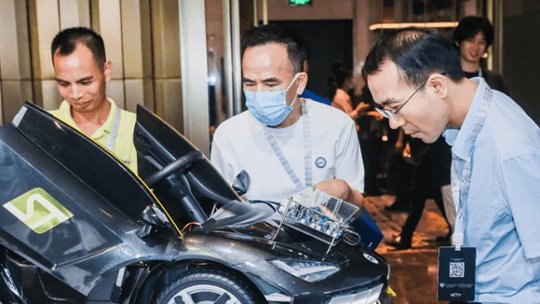 A group of men gathers around a sleek black sports car model in an indoor setting. The car features distinctive angular lines and a transparent section revealing internal components.
