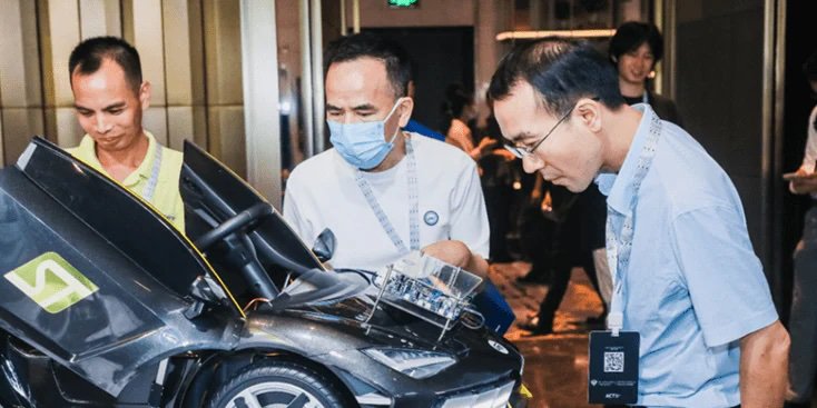 A group of men gathers around a sleek black sports car model in an indoor setting. The car features distinctive angular lines and a transparent section revealing internal components.