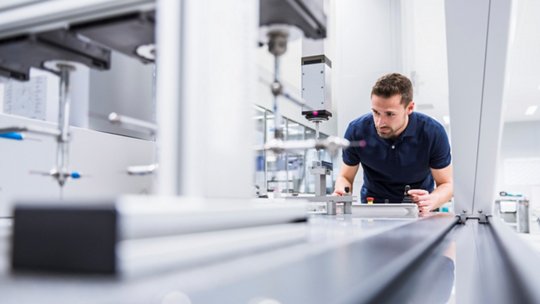 A technician or engineer leans over a long piece of industrial equipment in a bright, modern laboratory or manufacturing facility. 