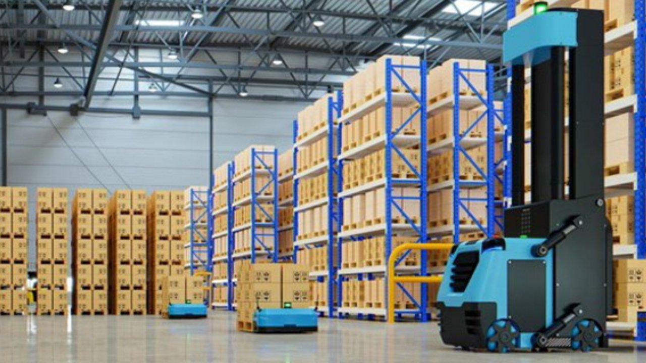 A quadcopter delivery drone flies through a warehouse or industrial yard carrying a cardboard box.