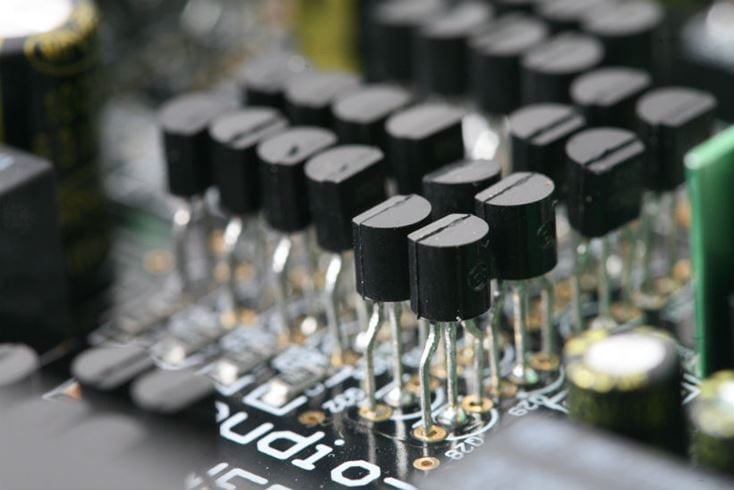 Multiple rows of large industrial batteries connected by thick black cables. The batteries are arranged in a clean, well-lit facility. Red and black connectors are visible on each battery.