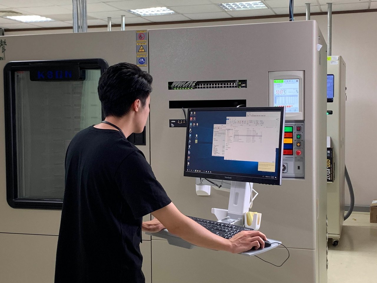 A young adult male technician stands at a workstation, interacting with a computer and control panel in a laboratory or industrial setting.