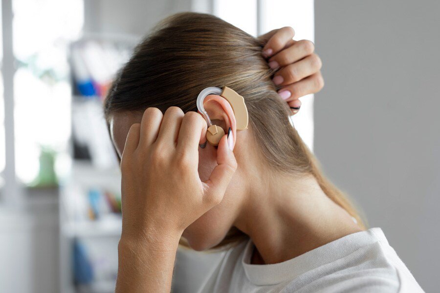 A woman is seen adjusting a beige hearing aid device behind her ear in a bright indoor setting.