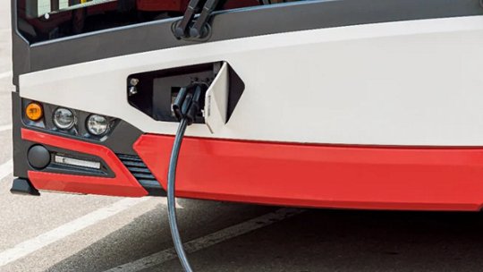 야외 주차장에 있는 충전 케이블에 연결된 전기 시내버스. 버스는 주차되어 있으며 차량 앞부분에 충전 포트가 뚜렷하게 보입니다.