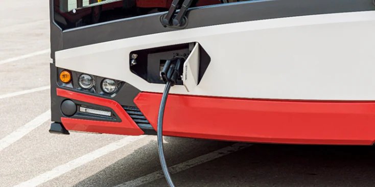 屋外の駐車場で充電ケーブルにつながれた電動シティバス。バスのフロント部分にある充電ポートがはっきりと見えています。