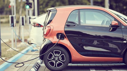 야외 주차장에 작은 투톤 전기차가 충전소에 연결되어 있으며, 근처에서 또 다른 흰색 차량이 충전 중입니다.