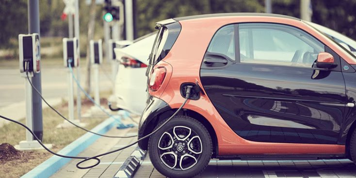 A small, two-tone electric car is plugged into a charging station in an outdoor parking area, with another white vehicle charging nearby.