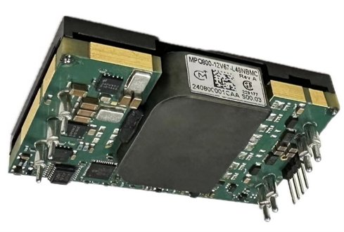 An isolated DC DC power converter module is shown in close-up against a plain white background. The green printed circuit board features multiple electronic components, including capacitors, resistors, and integrated circuits. A large black central block covers part of the board, while metal pins and connectors are visible on one side for electrical interfacing. A white identification label with technical markings and barcodes is attached to the top surface.