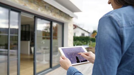 A woman stands outside a modern house, using a tablet device to interact with a smart home interface. The setting features large glass sliding doors and a contemporary exterior. The tablet screen displays graphs and controls, suggesting home automation or security monitoring. The overall mood is modern and tech-savvy, with natural daylight and a casual atmosphere.