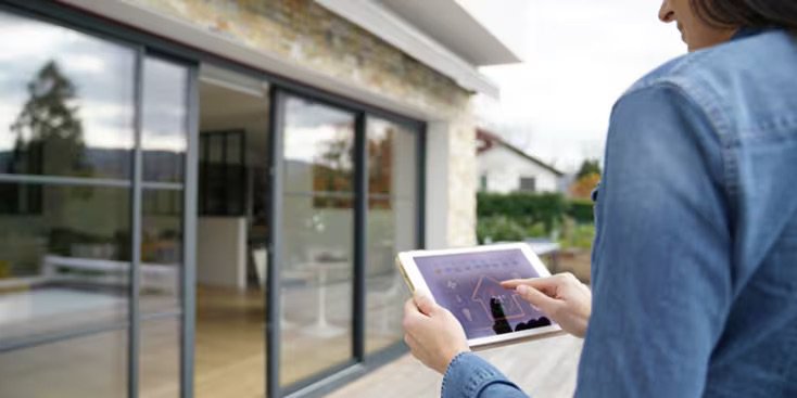 A woman stands outside a modern house, using a tablet device to interact with a smart home interface. The setting features large glass sliding doors and a contemporary exterior. The tablet screen displays graphs and controls, suggesting home automation or security monitoring. The overall mood is modern and tech-savvy, with natural daylight and a casual atmosphere.