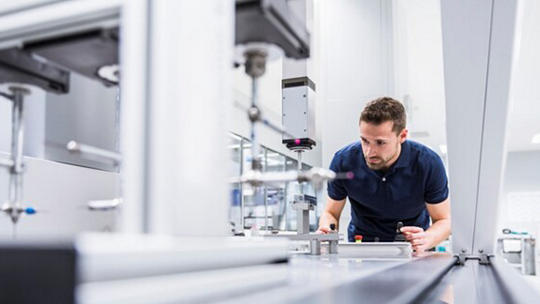 A person is closely examining a piece of automated machinery or equipment in a modern, brightly lit laboratory or industrial environment.