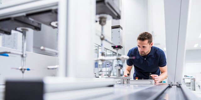 A person is closely examining a piece of automated machinery or equipment in a modern, brightly lit laboratory or industrial environment.