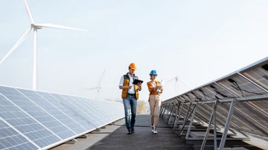 Two engineers wearing safety helmets and vests walk between rows of solar panels, conducting an inspection. The setting is an outdoor renewable energy facility with visible wind turbines in the background.