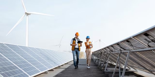 Two engineers wearing safety helmets and vests walk between rows of solar panels, conducting an inspection. The setting is an outdoor renewable energy facility with visible wind turbines in the background.
