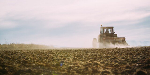 A tractor is seen working across a large, freshly plowed field under a bright sky. The scene captures the dust rising behind the vehicle as it moves through the soil.
