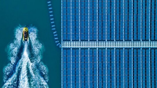 This aerial image captures a yellow boat moving through blue water, creating a wake as it passes beside a large array of floating solar panels. The solar panels are arranged in neat, geometric rows.